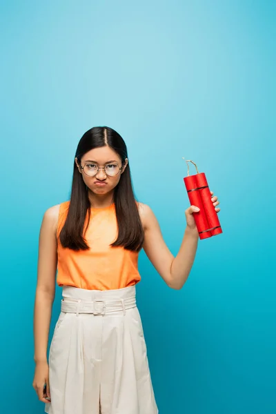 Dissatisfied asian girl in glasses holding dynamite sticks on blue — Stock Photo