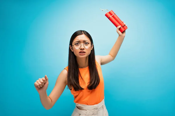 Emotional asian woman in glasses holding dynamite sticks above head on blue — Stock Photo