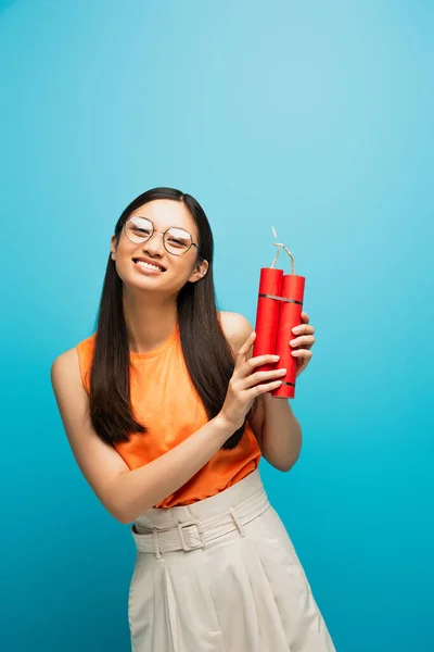 Happy asian girl in glasses holding dynamite sticks on blue — Stock Photo