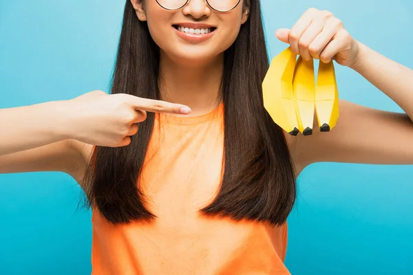 Abgeschnittene Ansicht eines fröhlichen Mädchens in Brille, das mit dem Finger auf reife Bananen zeigt, isoliert auf blauem Grund — Stockfoto
