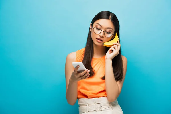 Chica asiática en gafas usando smartphone y sosteniendo plátano en azul - foto de stock