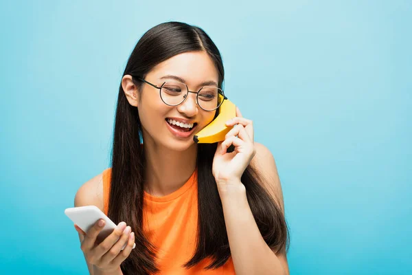 Felice ragazza asiatica in occhiali utilizzando smartphone e tenendo banana vicino all'orecchio su blu — Foto stock