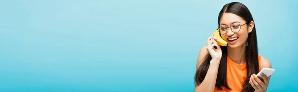 Panoramisches Konzept eines glücklichen asiatischen Mädchens in Brille mit Smartphone und Banane auf blau — Stockfoto