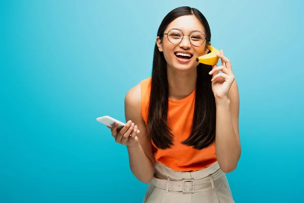 Sourire asiatique fille dans des lunettes en utilisant smartphone et tenant banane sur bleu — Photo de stock