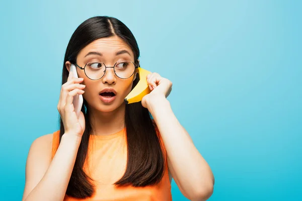 Surprised asian girl in glasses taking on smartphone and holding banana on blue — Stock Photo