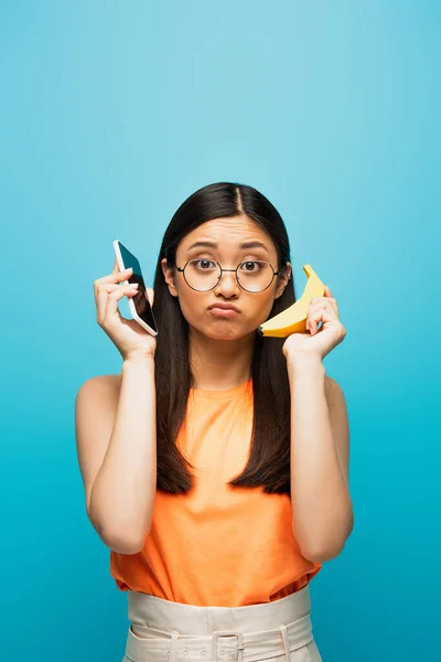 Confused asian girl in glasses holding smartphone and banana isolated on blue — Stock Photo