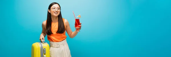 Concepto panorámico de chica asiática feliz en gafas celebración de cóctel y equipaje en azul - foto de stock