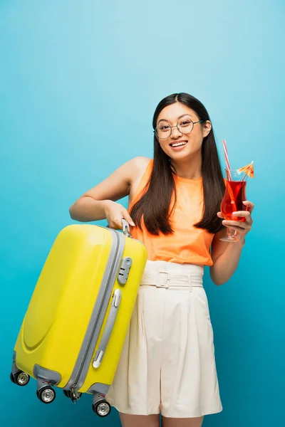 Alegre asiático menina no óculos segurando coquetel e bagagem no azul — Fotografia de Stock