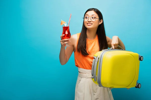 Alegre asiático menina no óculos segurando coquetel e bagagem enquanto olhando afastado no azul — Fotografia de Stock