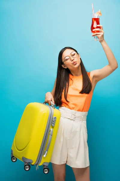 Весела азіатська жінка в окулярах тримає багаж і коктейль над головою на синьому — стокове фото