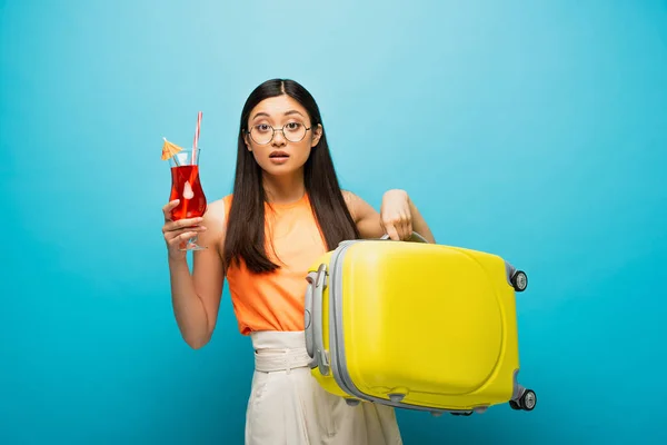 Junge asiatische Frau in Brille mit Gepäck und Cocktail auf blau — Stockfoto