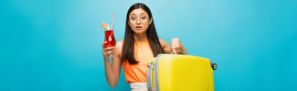 Panoramic concept of pretty asian woman in glasses holding baggage and cocktail on blue — Stock Photo