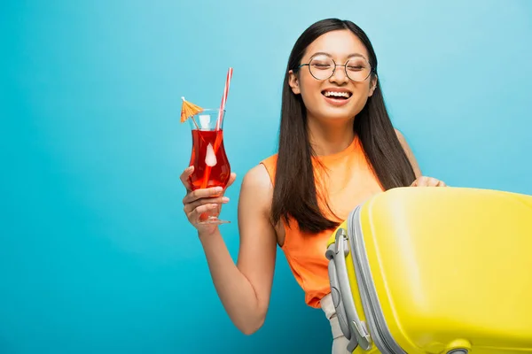 Весела азіатська жінка в окулярах тримає коктейль і багаж ізольовані на синьому — стокове фото