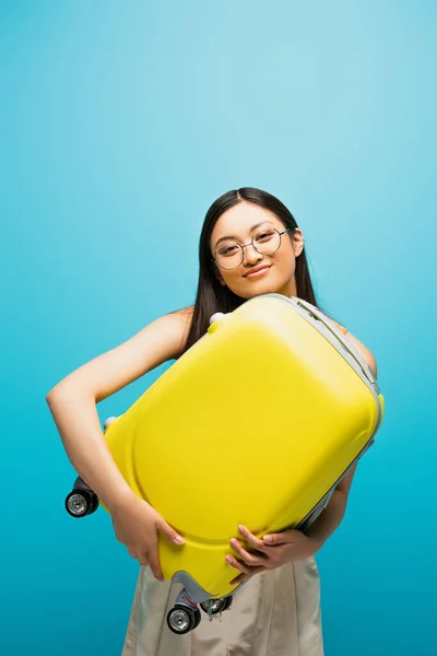 Cheerful asian woman in glasses holding yellow luggage on blue — Stock Photo