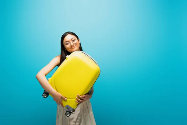 Positivo asiático mujer en gafas celebración de amarillo equipaje en azul - foto de stock
