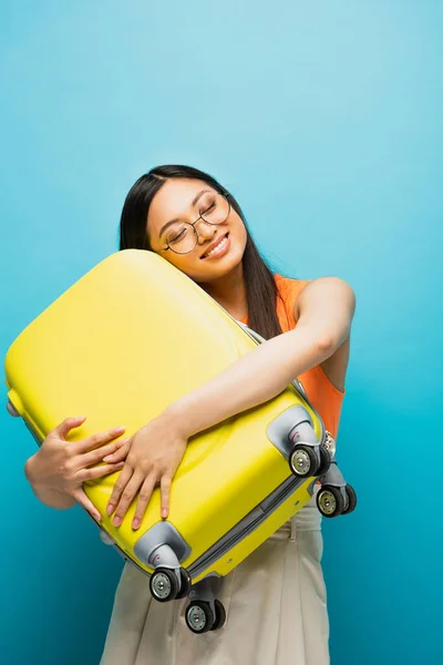Весела азіатська жінка в окулярах обіймає жовтий багаж на синьому — стокове фото