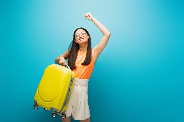 Весела азіатська жінка в окулярах тримає жовтий багаж і жестикулює на синьому — стокове фото