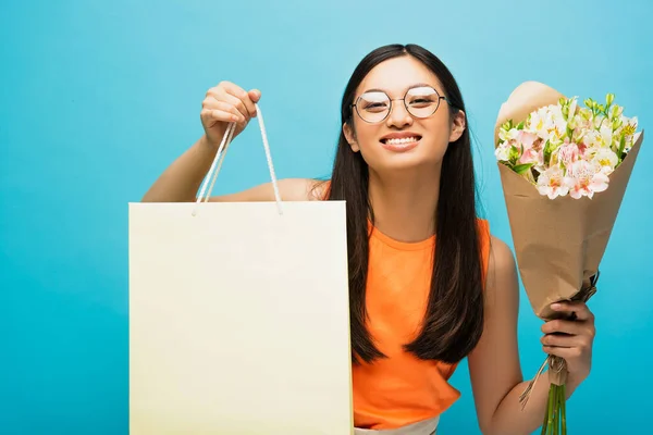 Positive asian woman in glasses holding shopping bag and flowers on blue — Stock Photo