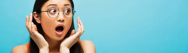 Horizontal crop of shocked asian woman in glasses gesturing and looking away isolated on blue — Stock Photo