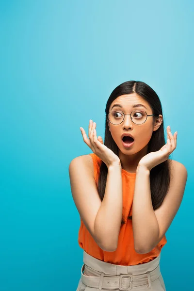 Sorprendido asiático mujer en gafas gesto y mirando lejos aislado en azul - foto de stock