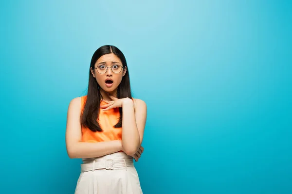 Sorprendida mujer asiática en gafas de pie en azul - foto de stock