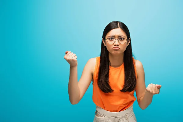 Frustrated asian girl in glasses showing clenched fists on blue — Stock Photo