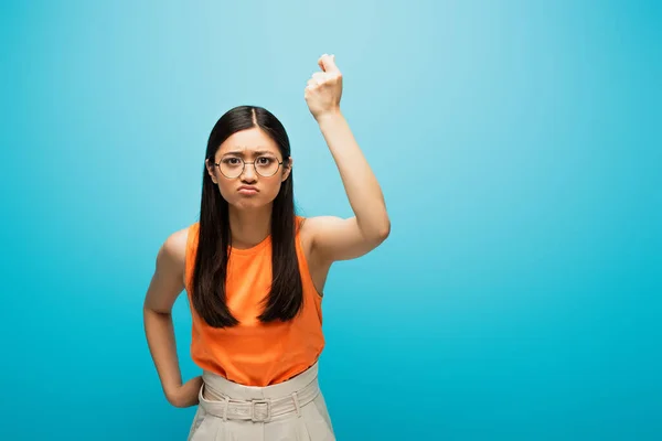 Frustré asiatique fille dans lunettes montrant serré poing sur bleu — Photo de stock