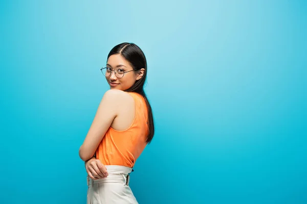 Позитивная азиатская девушка в очках улыбается стоя на синем — стоковое фото