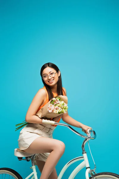 Cheerful asian girl in glasses riding bicycle and holding flowers on blue — Stock Photo