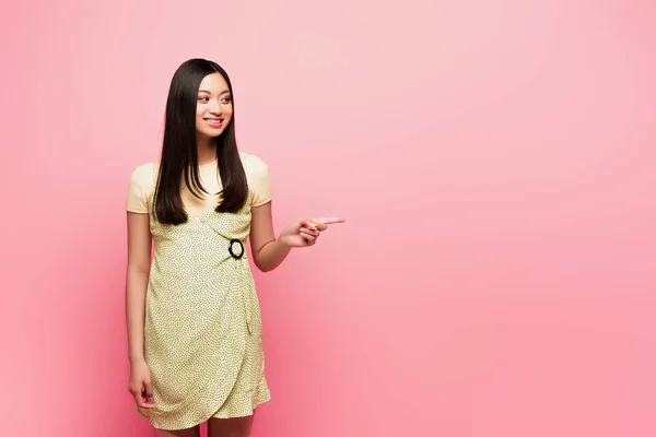 Cheerful asian girl pointing with finger and looking away on pink — Stock Photo