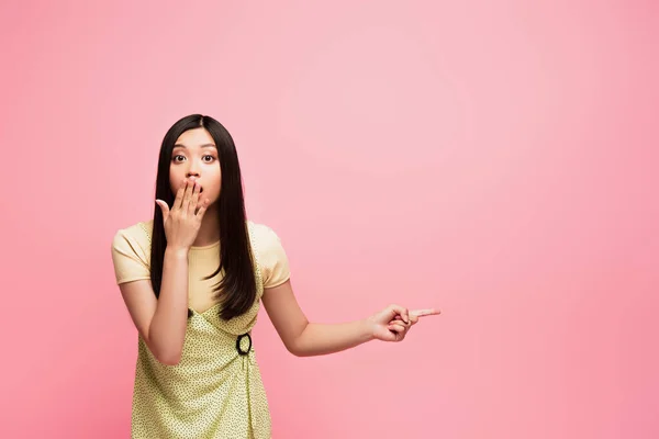 Conmocionada chica asiática señalando con el dedo y cubriendo la boca aislado en rosa - foto de stock