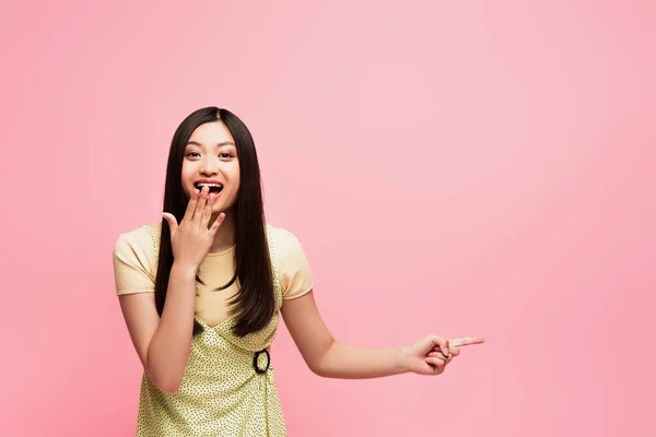 Excited asian girl pointing with finger and covering mouth isolated on pink — Stock Photo