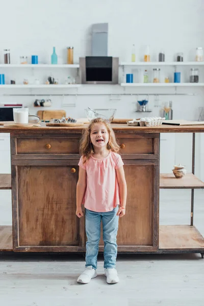 Full lengtn vista da menina de pé perto da mesa da cozinha com ingredientes e utensílios de cozinha — Fotografia de Stock