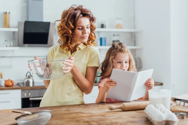 Selective focus of girl reading cookbook near mother holding glass bowl with whisk — Stock Photo