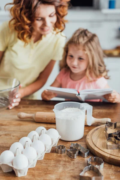 Selective focus of chicken eggs, flour in measuring jug and cookie cutters near mother and child reading cookbook — Stock Photo