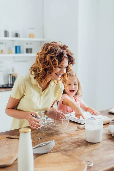 Fuoco selettivo del bambino che punta con il dito al libro di cucina vicino alla madre che tiene la ciotola di vetro con la frusta — Foto stock