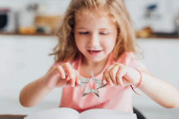 Foyer sélectif de fille blonde tenant coupe-biscuits en forme d'étoile tout en lisant le livre de cuisine — Photo de stock