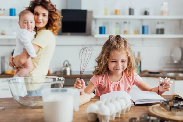 Enfoque selectivo del niño sosteniendo batidor y leyendo libro de cocina cerca de la mesa con utensilios de cocina, y la madre joven sosteniendo al bebé sobre el fondo — Stock Photo