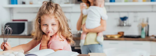 Messa a fuoco selettiva della ragazza premurosa che tiene frusta e legge il libro di cucina vicino alla madre con bambino sullo sfondo, concetto panoramico — Foto stock