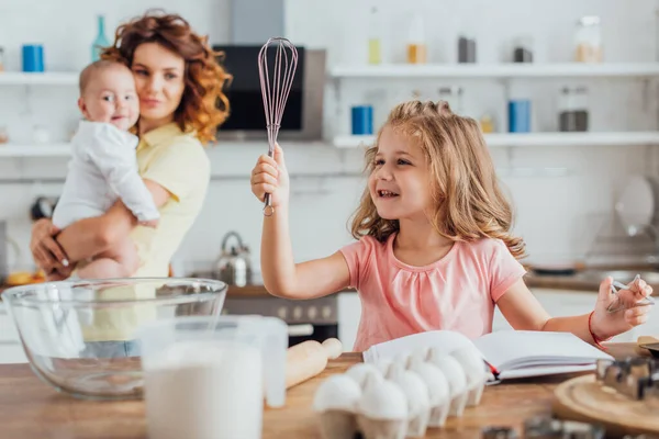 Foco seletivo da criança segurando batedor perto da mesa com ingredientes e utensílios de cozinha, e mãe com bebê no fundo — Fotografia de Stock