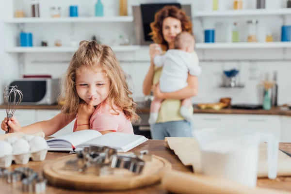 Messa a fuoco selettiva della ragazza che tiene frusta e legge il libro di cucina vicino alla madre che tiene il piccolo figlio — Foto stock