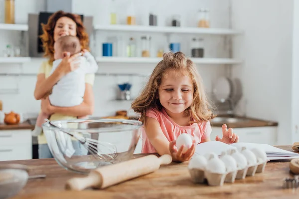 Selective focus of girl holding chicken egg near bowl, whisk and rolling pin near mother with little son — Stock Photo
