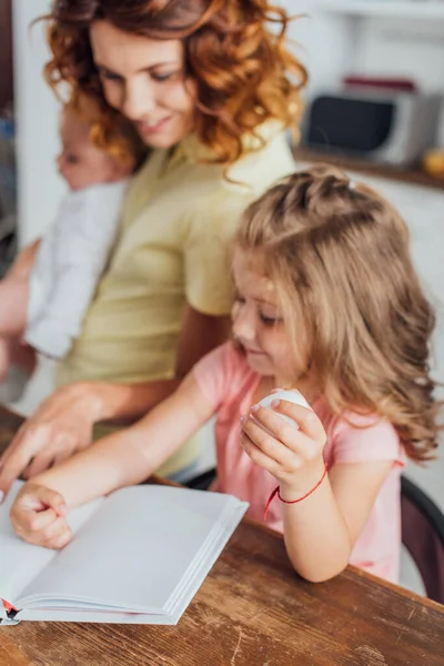 Selektiver Fokus der Mutter, die mit dem Finger auf Kochbuch zeigt, während Tochter Hühnerei hält — Stockfoto