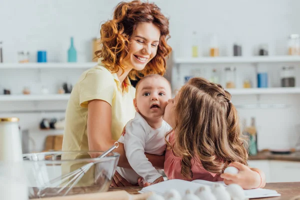 Enfoque selectivo de la madre joven sosteniendo hijo bebé mientras hija besándolo y sosteniendo huevo de gallina en la cocina - foto de stock