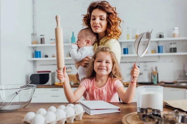 Messa a fuoco selettiva della ragazza che tiene il mattarello e setaccio vicino alla giovane madre con bambino in cucina — Foto stock