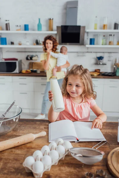 Selective focus of child pointing with finger at cookbook and holding bottle of milk near mother with infant son — Stock Photo