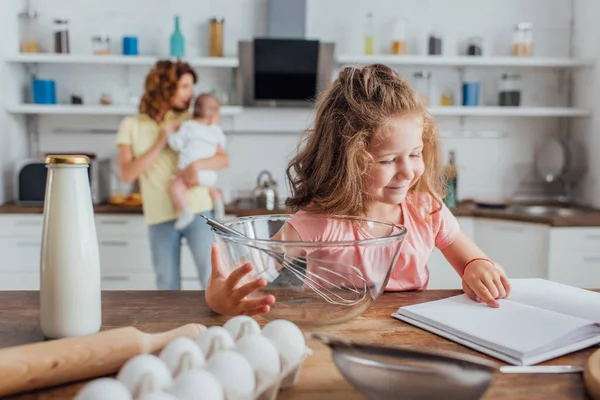 Selective focus girl pointing with finger at cookbook near bowl, whisk, bottle of milk and chicken eggs — Stock Photo