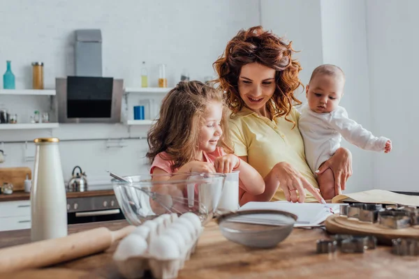 Selective focus of young mother holding son and pointing with finger at cookbook near daughter, cooking utensils and ingredients on table — Stock Photo