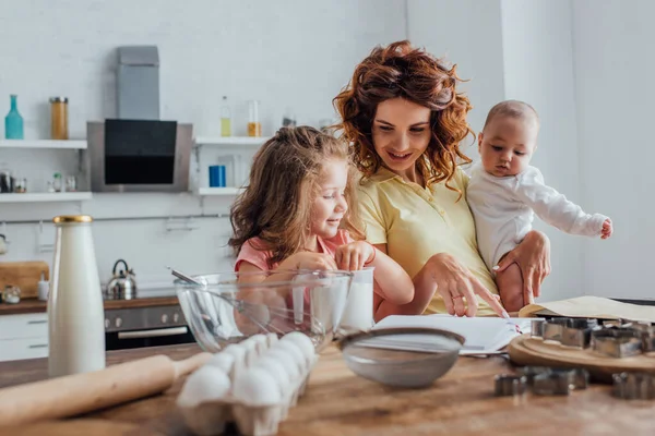 Foco seletivo da jovem mãe segurando filho e apontando com o dedo para o livro de receitas perto da filha, utensílios de cozinha e ingredientes na mesa — Fotografia de Stock