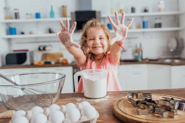 Messa a fuoco selettiva della ragazza bionda mostrando le mani nella farina vicino al tavolo della cucina con farina, uova, tagliabiscotti e frusta in ciotola — Foto stock